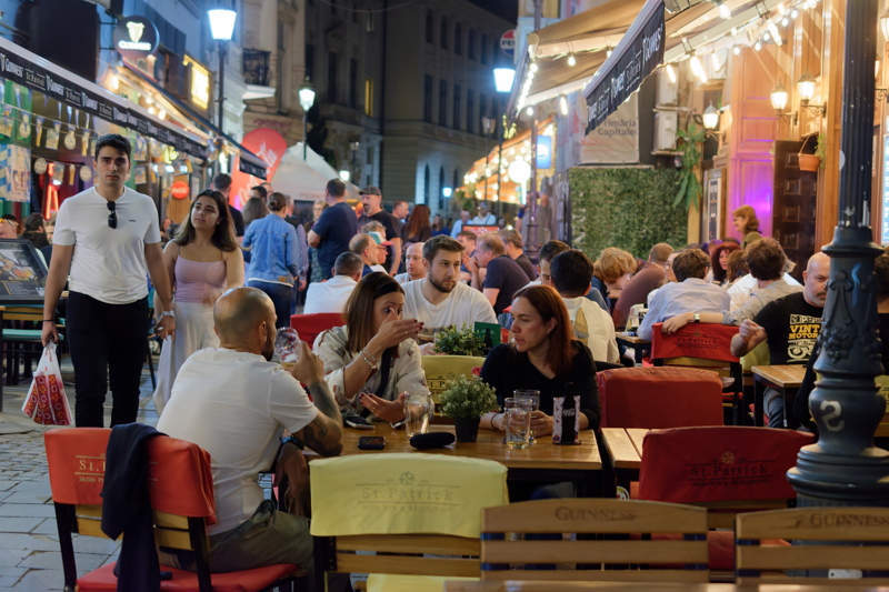 18 - A lively outdoor terrace scene at night in the Old Town of Bucharest, Romania, with people sitting at tables and walking along the cobblestone pedestrian street